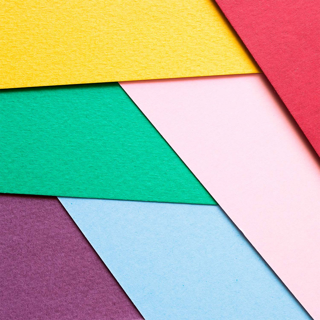 A close-up arrangement of overlapping colored paper or fabric swatches in yellow, red, green, pink, light blue, and purple, arranged in diagonal geometric shapes.