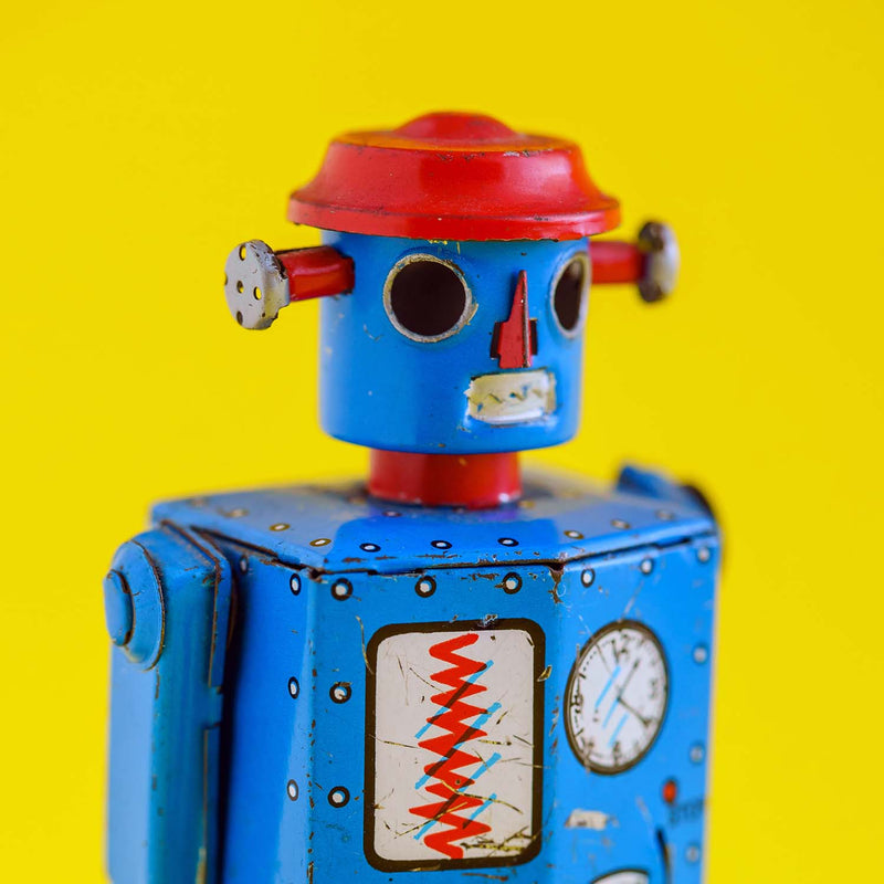 A vintage tin toy robot with a blue body, red dome-shaped head, and silver metal joints, set against a solid bright yellow background.