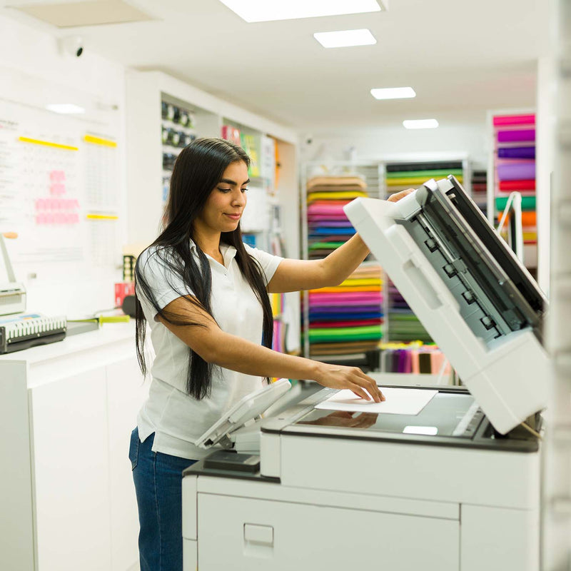 A woman in a white t-shirt and blue jeans operates a white and black office photocopier in a brightly lit office supply shop.