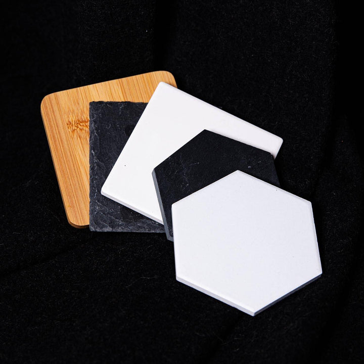Three square coasters with rounded corners displayed in a fan arrangement on a black textured background. The set includes a light natural bamboo coaster, a dark charcoal slate coaster, and two glossy white ceramic hexagonal coasters.