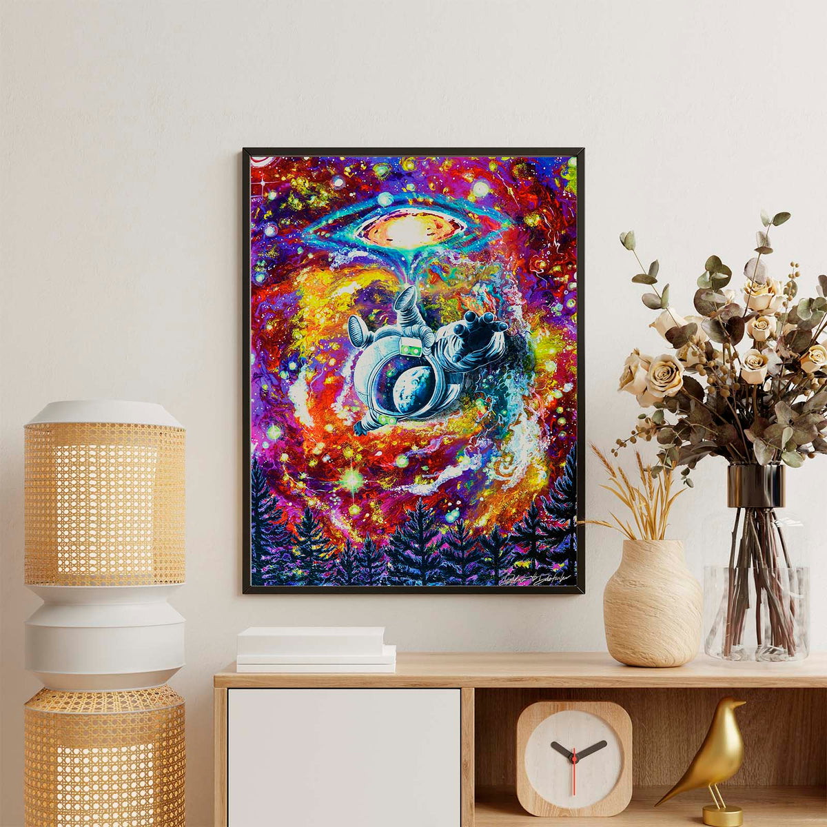 A colorful framed wall art print featuring a cosmic space scene with a UFO and astronaut, displayed on a light wall above a wooden console table with decorative lamps, vases, and a clock.