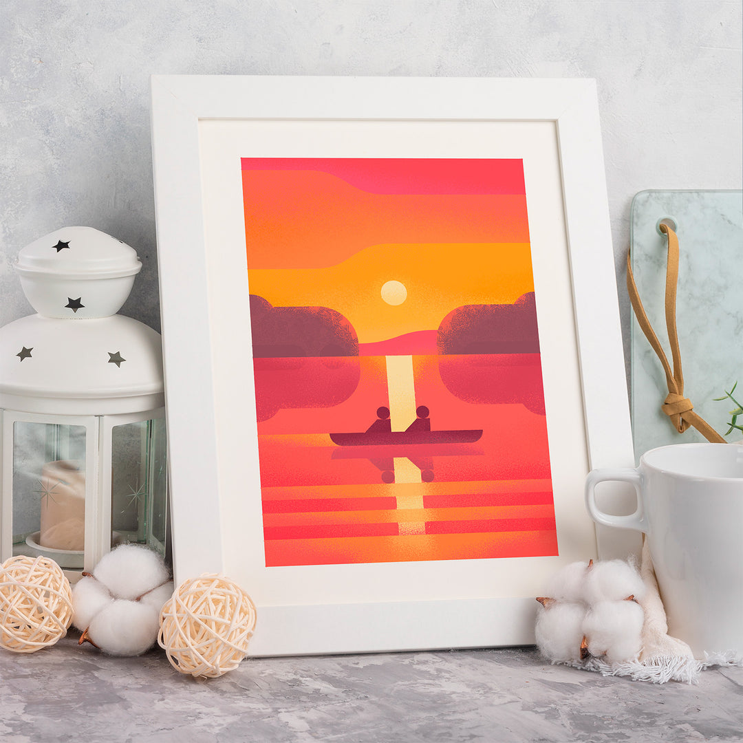 Framed artwork of a sunset over water with two figures in a boat, placed on a surface with decorative items.